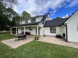 White farmhouse with installed gutters, dark roof, rear patio, and manicured lawn